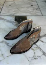 Bota corta de cuero color marrón, con taco cuadrado y punta fina. Presenta una correa con tachas pequeñas y una argolla metálica decorativa alrededor del tobillo. Cuenta con cierre lateral.