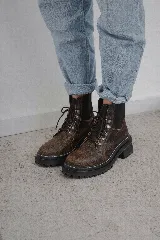 Borcego de gamuza color chocolate con paneles laterales elásticos negros y suela track dentada. Presenta sistema de cordones con ganchos metálicos y tirador en la parte trasera.