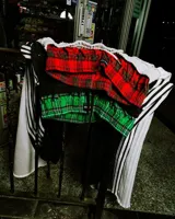 Pantalón deportivo negro de corte ancho, con tres franjas blancas verticales a lo largo de las piernas. La cintura es elástica y presenta un forro interior con estampado a cuadros en tonos verde y negro.