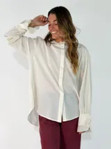 Camisa de algodón color blanco, con cuello redondo y detalle de volados en el cuello y puños. Presenta mangas largas con puños elásticos y un diseño de corte holgado con ruedo asimétrico, más largo en la parte trasera.