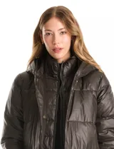 Campera puffer larga de color marrón oscuro, con diseño capitoneado de costuras horizontales. Presenta un sistema de cierre doble con pechera interna fija con cierre y cuello alto, y cierre frontal exterior con botones a presión. Incluye bolsillos frontales amplios, capucha fija y cierres laterales en el ruedo para mayor movilidad.