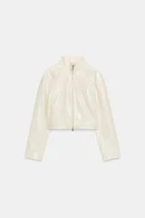 Chaqueta corta de efecto piel en color crema, con cuello subido, manga larga y cierre frontal con cremallera. Presenta bolsillos de vivo en el pecho y en la parte delantera.