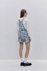 Jumper de jean corto, de color celeste claro con estampado floral en tonos oscuros. Presenta tirantes regulables con hebillas metálicas, bolsillo pectoral y bolsillos laterales funcionales.