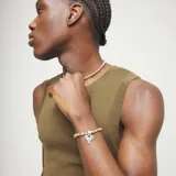 Pulsera de eslabones tipo bola de metal bañada en plata, con un dije central con forma de corazón irregular y un cierre de placa rectangular. Incluye un pequeño dije con la inscripción "UNO & 50".