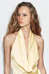 Vestido mini satinado de color amarillo pastel, con cuello halter, escote drapeado y detalle de fajín drapeado con lazo lateral. Cuenta con forro interior y cierre lateral con cremallera oculta.