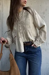 Camisa beige de manga larga con detalles bordados en el frente, cuello redondo y cierre frontal con botones. Las mangas presentan puños con volados.
