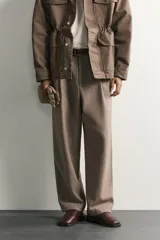 Pantalón relaxed fit de hombre color marrón con textura estructurada, cintura con pliegues frontales, bolsillos laterales y cierre frontal con cremallera y botón.