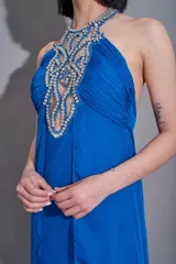 Vestido largo de gasa color azul royal, sin mangas, con escote halter y espalda descubierta en forma de V. Presenta drapeado en la falda y bordado en el escote.