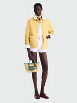 Cartera tipo canasta de Longchamp, modelo Le Panier Pliage XS, confeccionada en paja trenzada con detalles en cuero verde. Presenta asas cortas, correa ajustable y un dije decorativo con cuentas de madera.