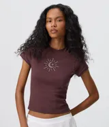 Remera baby tee de algodón color marrón oscuro, con cuello redondo, manga corta y calce ajustado. Presenta un aplique frontal de pedrería brillante con diseño místico de sol, luna y estrella.