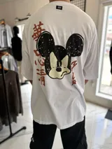Remera oversize blanca de manga corta, con cuello redondo y etiqueta decorativa en la parte posterior del cuello. Presenta un gran estampado en la espalda que combina un diseño estilizado de Mickey Mouse con caracteres en estilo asiático en color rojo.
