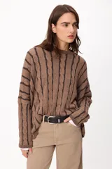 Sweater de punto con diseño de trenzas verticales, cuello redondo y corte crop. Presenta una combinación de colores crudo y camel, con mangas largas y ajuste holgado.