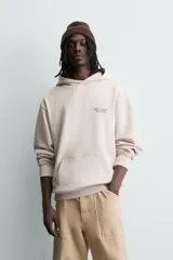Sudadera hoodie relaxed fit de color beige, con cuello con capucha, manga larga y bolsillo frontal tipo canguro. Presenta un pequeño estampado de texto en contraste en el pecho.