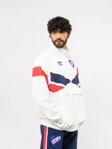 Buzo deportivo de nylon con medio cierre frontal y cuello alto. Presenta un diseño de bloques de color en azul, blanco y bordó, con el escudo del Club Nacional de Football y el logo de Umbro en el pecho.