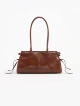 Cartera Kate de cuero vacuno diseñada para llevar al hombro, con cierre principal y bolsillo interno. Presenta aplique de candado y llavero tipo birkin en el frente, además de tiras laterales decorativas.