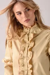 Camisa celeste con volados decorativos en los hombros, pecho, mangas y ruedo, destacados con ribetes en tono rojo. Presenta cierre frontal de botones, cuello alto y mangas largas abullonadas con puños ajustados.