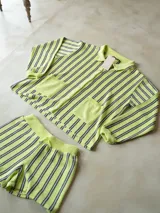 Conjunto de dos piezas de tejido de punto a rayas verticales en tonos verde lima, gris y blanco. Incluye una camisa de manga larga con cuello polo, cierre frontal de botones y dos bolsillos delanteros, junto con un short de tiro alto con cintura elástica y cordón ajustable.