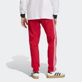 Pantalón deportivo Adidas de corte ajustado, color rojo con las icónicas tres tiras blancas laterales y logo del trébol bordado en la parte superior. Confeccionado en tejido de piqué con cintura elástica y cordón de ajuste.