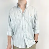 Camisa celeste con rayas verticales blancas, grises y beige. Tiene cuello italiano, manga 3/4 y corte semi entallado.