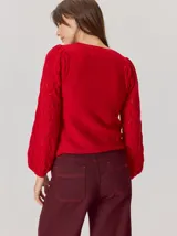 Buzo de lana color beige con mangas abullonadas de tejido calado con diseño de rombos. Presenta un cuello redondo y un detalle de flor negra de tela con cintas colgantes en el escote.