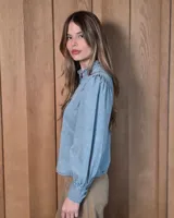 Camisa de jean blanca con cuello alto con volados, cierre frontal con botones oscuros y mangas largas abullonadas con puños ajustados.