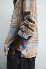 Camisa de manga larga con cuello solapa y dos bolsillos frontales de plastrón con botón. Presenta un estampado de cuadros en tonos beige, azul claro y marrón.