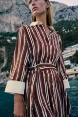 Vestido midi camisero de rayas verticales en tonos bordó y blanco, con cuello solapa y puños en color contrastante. Cuenta con mangas largas, cierre frontal de botones y un cinturón ajustable con hebilla metálica en la cintura.