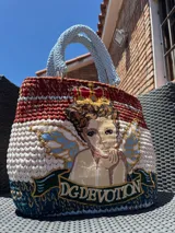 Bolso tejido a mano con hilo de trapillo, eco-cuero y textiles, con un diseño central de querubín coronado y alas, sobre un fondo de franjas rojas, blancas y azules. Posee asas cortas tejidas en color celeste claro.