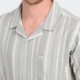 Camisa de manga corta con cuello cubano, color gris claro con rayas verticales en blanco y gris más oscuro. Presenta un bolsillo en el pecho y cierre frontal de botones.