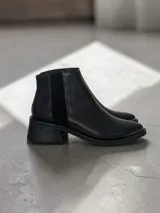 Bota corta de cuero negro con diseño tipo chelsea, presenta un panel elástico lateral y un taco cuadrado robusto. Incluye un detalle metálico en la puntera.