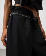 Pantalón negro de corte ancho y tiro alto con diseño asimétrico. Presenta un cruce frontal con cinturón de tela a cuadros grises y negros, que deja ver un forro o panel interior de la misma tela a cuadros.