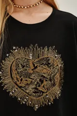 Remera negra de corte holgado con cuello redondo y mangas cortas. Presenta un aplique central en forma de corazón bordado con hilos y cuentas doradas, con un diseño detallado de aves en su interior.