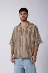Camisa de manga corta con cuello solapa, corte holgado y cierre frontal de botones. Presenta un diseño de rayas verticales en tonos beige, negro y blanco.