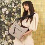 Cartera bandolera gris de formato amplio, con correa larga regulable y desmontable con inscripciones. Cuenta con cierre superior, bolsillo frontal con cierre y detalle colgante de cordones, y un bolsillo trasero con cierre.