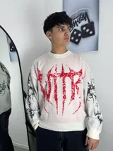 Sweater de punto color crema con diseño gráfico estilo metal en color rojo en el frente y detalles estampados en negro en las mangas. Presenta cuello redondo y puños acanalados.