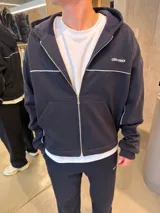 Campera con capucha negra de corte boxy, con cierre frontal completo, bolsillos tipo canguro y detalles de líneas blancas horizontales y en las mangas. Cuenta con un pequeño logo bordado en el pecho.