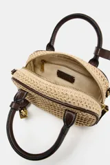 Bolso pequeño tipo bowling de rafia tejida en color natural, con ribetes, asas de mano y detalles en cuero marrón oscuro. Incluye un charm colgante con candado dorado y una correa ajustable extraíble para llevar al hombro o cruzado.