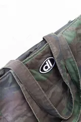 Bolso tote de tela con estampado de camuflaje en tonos verdes y marrones. Tiene asas de hombro y un logo ovalado con letras blancas en el centro.
