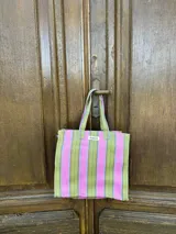 Bolso tipo tote bag con estampado a rayas verticales en tonos verde, rosa, celeste y marrón. Tiene asas de hombro y un parche con la inscripción "AMÉN".