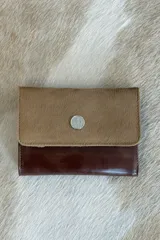 Billetera de diseño bicolor, con solapa superior en cuero con pelo color tostado y cuerpo inferior en cuero liso color marrón oscuro. Presenta un aplique metálico circular plateado con logo grabado en el centro.