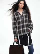 Camisa de corte relajado con estampado de cuadros en tonos negro y beige. Presenta cuello camisero, cierre frontal parcial con botones y mangas largas con puños ajustables mediante lazos. El diseño destaca por un detalle de encaje en la espalda que aporta contraste.