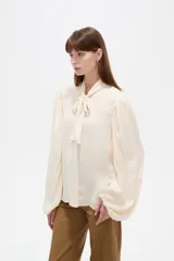 Camisa de poliéster color marfil, de diseño holgado, con cuello alto lazada y mangas largas abullonadas.