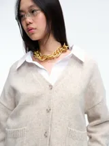 Cardigan de punto color beige con cierre frontal de botones, cuello en V y dos bolsillos delanteros. Presenta mangas largas con puños acanalados y un diseño de corte holgado.