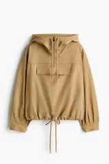 Campera popover color tostado confeccionada en sarga de mezcla de algodón y lyocell. Cuenta con capucha con cordón ajustable, medio cierre frontal y un amplio bolsillo delantero con solapa. Presenta diseño de hombros caídos, canesú frontal, mangas abullonadas con pliegues, puños con cierre ajustable de botón y bajo con cordón interior para fruncir.