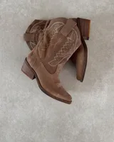 Bota corta de gamuza color marrón topo, estilo western, con bordados decorativos en hilo blanco en caña y empeine. Posee taco cuadrado de madera y punta ligeramente afilada.