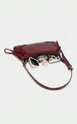 Cartera tipo baguette de charol color bordó, con diseño estructurado, cierre superior y detalles de tachas metálicas en las esquinas inferiores. Incluye tiras laterales decorativas y correa de hombro ajustable.