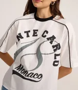 Remera blanca de algodón con cuello redondo y ribete negro. Presenta un estampado grande con el texto "MONTE CARLO" y "Monaco" en tonos negro y gris, con un diseño que simula un número 8 o un símbolo de infinito.