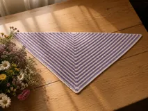 Chal triangular tejido a mano con diseño de rayas horizontales en tonos lila y blanco. Presenta una textura de punto fino y un borde tejido en color blanco.