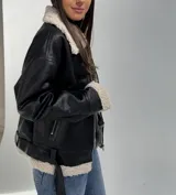 Campera tipo aviador de cuero sintético negro con interior y detalles de corderito en color crema. Presenta cierre frontal, bolsillos laterales con cierre y cinturón ajustable en el ruedo.