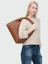 Cartera tipo hobo Marc Jacobs modelo The Sack, confeccionada en cuero granulado color marrón. Presenta un diseño minimalista con el nombre de la marca y el modelo estampados en el frente y una correa integrada al cuerpo de la cartera.
